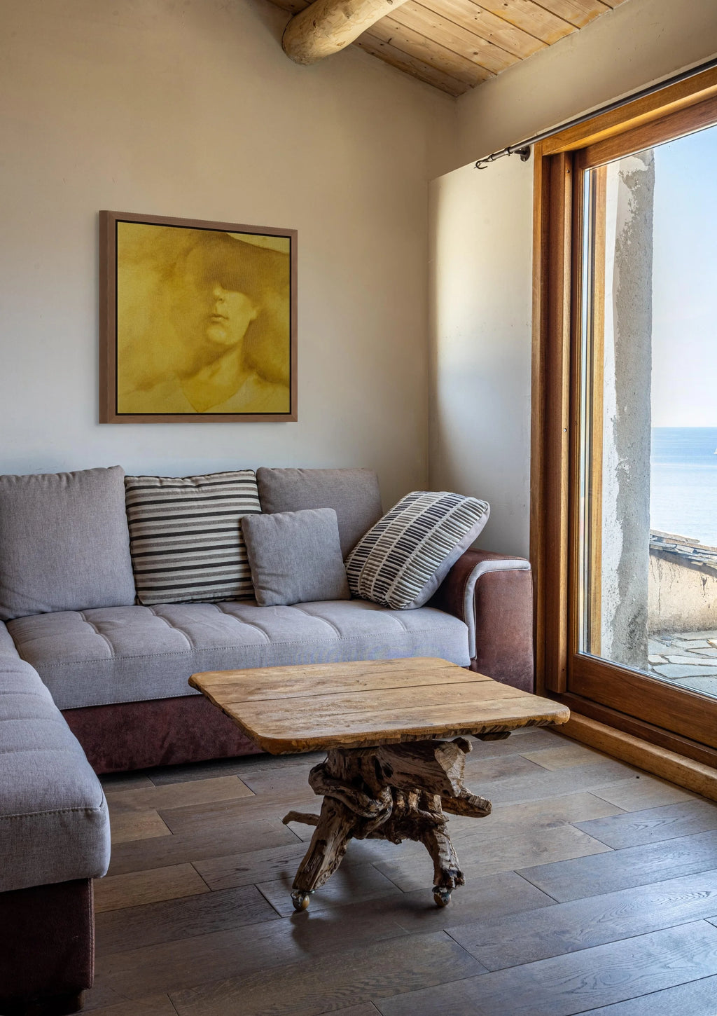 A styled interior living room corner featuring a light gray sectional sofa with striped and textured pillows, a rustic wood coffee table, a framed yellow portrait painting on the wall, and a large glass door looking out to the sea.