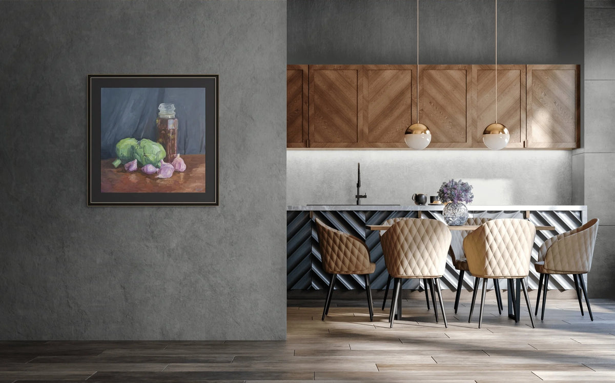 A framed still life painting of green artichokes and a jar hangs on a textured gray wall in a modern kitchen setting with wooden cabinets, pendant lights, and a dining table with beige chairs.