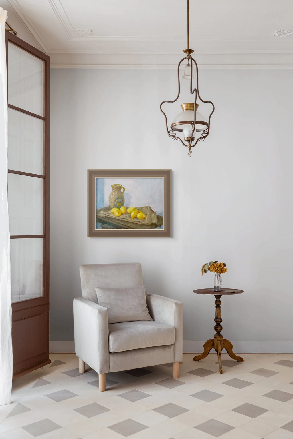 A styled interior room scene featuring a light beige armchair, a small round wooden side table with a vase of yellow flowers, a framed still life painting on the wall, and a hanging brass pendant light over a checkered tile floor.