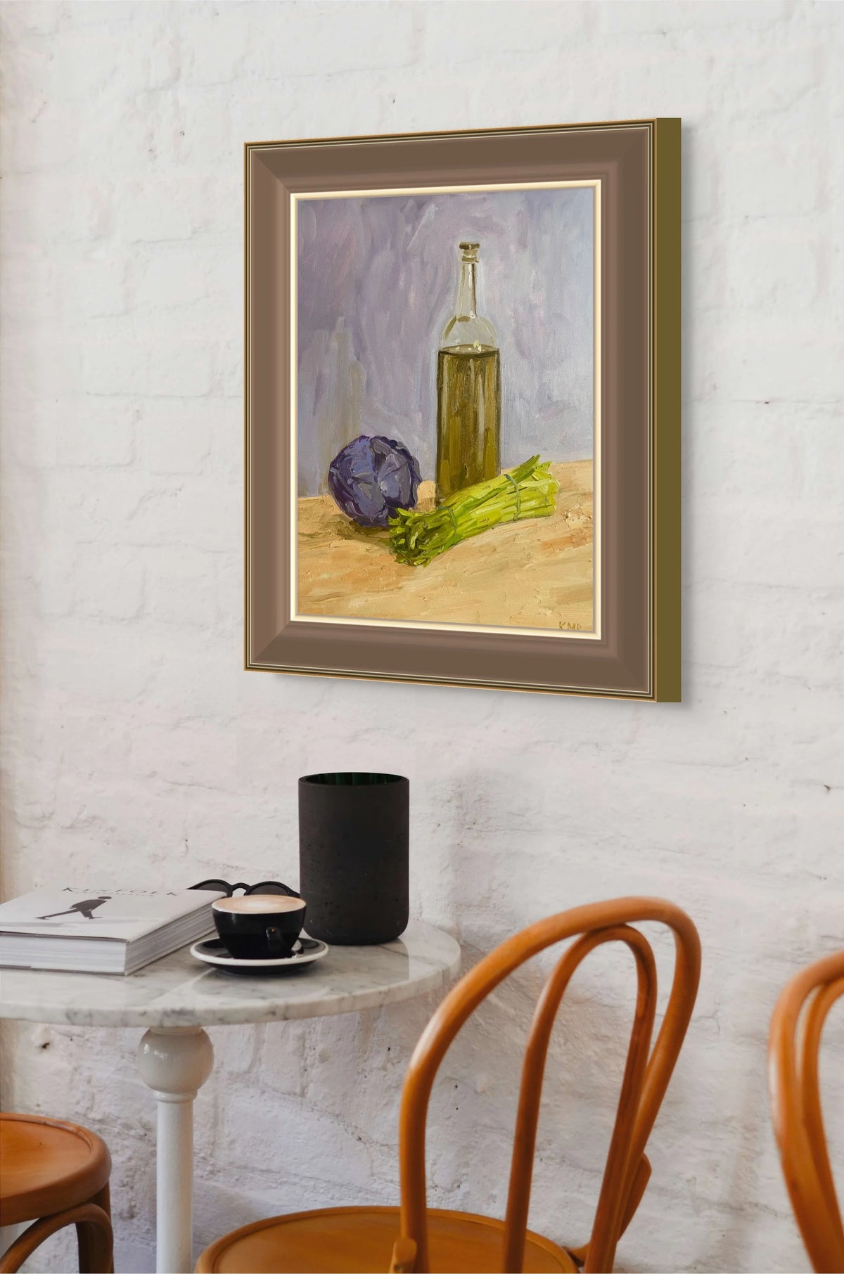 A framed still life painting hanging on a white brick wall above a small table with chairs. The painting depicts a glass bottle of olive oil, a purple cabbage and green asparagus on a yellow surface against a soft gray background. 